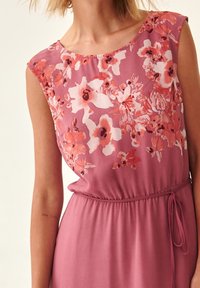 Robe florale en tissu rose doux avec de courtes manches capes, dotée d'une taille cintrée et d'un cordon de serrage. Les détails incluent des fleurs roses et blanches vives.