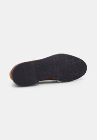 Suola di scarpa in suede marrone con una sezione in gomma nera texturizzata. Presenta un motivo di piccole perforazioni e un design della punta arrotondata.