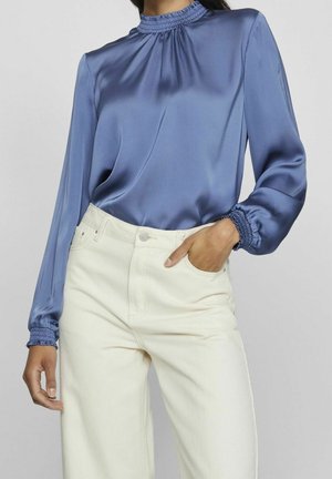 Kobieta w niebieskiej satynowej bluzce z długim rękawem z falbankami przy mankietach i jasnych, kremowych spodniach z wysokim stanem, z jedną ręką w kieszeni.