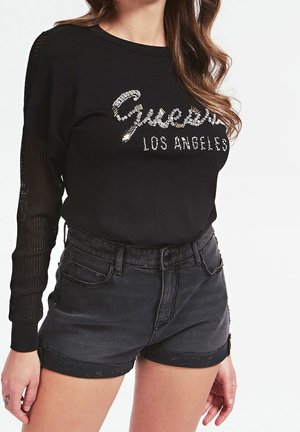 Mujer con un suéter negro con "Guess Los Angeles" en pedrería y shorts vaqueros negros de talle alto con dobladillos enrollados.