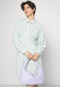 Vestido camponês em xadrez verde claro com colarinho e mangas compridas. Uma bolsa de ombro suave em lavanda é segurada à frente.