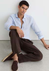 Mężczyzna z kręconymi włosami, ubrany w jasnoniebieską koszulę, ciemnobrązowe spodnie i pasujące mokasyny, siedzący na podłodze przy białej ścianie.