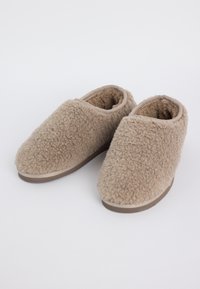 Een paar beige pluizige binnenpantoffels met een zachte textuur en bruine rubberen zolen, getoond op een witte achtergrond.