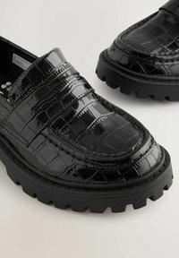 Czarne loafers z błyszczącą, wytłaczaną teksturą krokodyla, z masywną czarną gumową podeszwą i szczegółowym przeszyciem wzdłuż krawędzi.