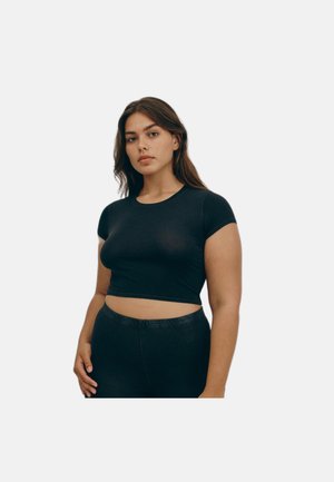 Joven mujer con cabello largo y oscuro, vistiendo un top corto negro ajustado y pantalones negros contra un fondo claro y liso.