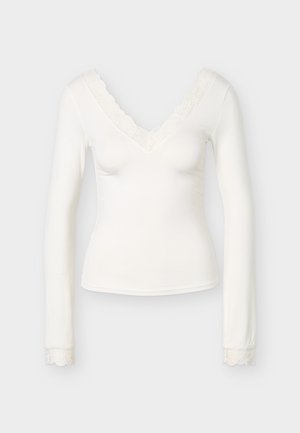 Balta ilgomis rankovėmis moteriška palaidinė su V formos iškirpte ir nėrinių apdaila ant apykaklės bei rankogalių, aptemptos silueto, pateikta ant vienspalvio fono.