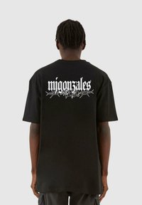 Svart oversized t-skjorte med et hvitt grafisk trykk av "migon sales" og blomsterdetaljer på ryggen. Korte ermer, rund hals, laget av bomull.