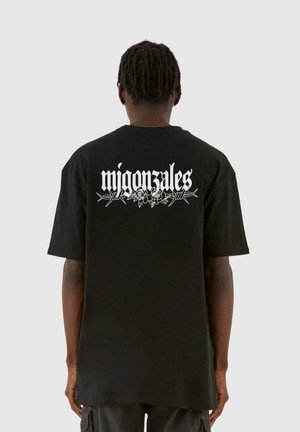 Svart oversized t-skjorte med et hvitt grafisk trykk av "migon sales" og blomsterdetaljer på ryggen. Korte ermer, rund hals, laget av bomull.