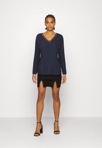 Top de mangas longas azul-marinho com decote em V e detalhe em costura laranja, combinado com uma saia mini preta com abertura frontal e um design ajustado.