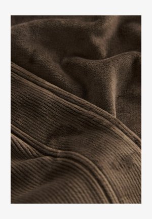 Brauner Stoff mit einer weichen, strukturierten Oberfläche, der sowohl glatte als auch gerippte Abschnitte aufweist. Das Material wirkt plüschig und langlebig.