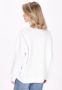 Felpa bianca con vestibilità ampia, polsini e orlo a costine, caratterizzata da un collo rotondo e una texture morbida. Indossata con jeans azzurri chiari.