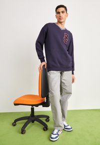 Jūras spēku džemperis ar V veida kakla izgriezumu un sarkanu izšūtu logo, pelēkas bikses un zilas sporta kurpes. Blakus modelim ir oranža biroja krēsls.