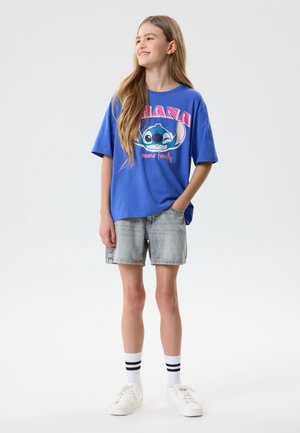 Jente som står og har på seg en blå, stor T-skjorte med Stitch-trykk, falmede denimshorts, hvite sokker med svarte striper og hvite joggesko.