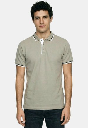 Homme portant un polo à manches courtes gris clair avec un col et des bords de manches rayés noir et blanc, debout devant un fond uni.