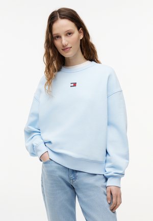 Junge Frau mit langen, welligen Haaren, die einen hellblauen Tommy Jeans Sweatshirt und hellblaue Jeans trägt, steht mit einer Hand in der Tasche.