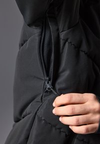 Jachetă puffer neagră cu țesătură texturată, cusături vizibile și un fermoar cu maner, parțial desfăcut pentru a dezvălui interiorul.