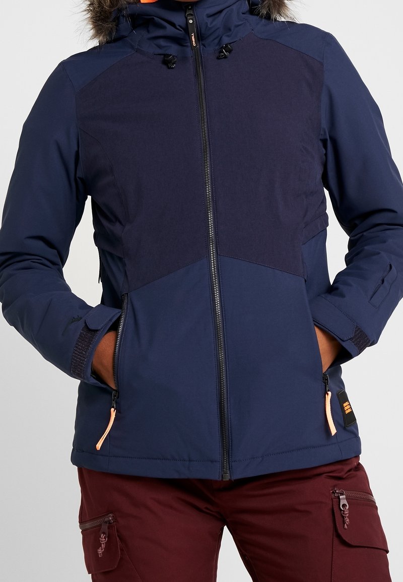 Mężczyzna w granatowej zimowej kurtce z futrzanym kapturem i bordowych spodniach, ręce w kieszeniach kurtki, zamek błyskawiczny z przodu zamknięty.