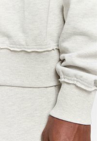 Hellgrauer Sweatshirt mit gerippten Bündchen und Saum. Der Stoff wirkt weich mit einer leicht strukturierten Oberfläche und einem ausgefransten Kanten-Detail.