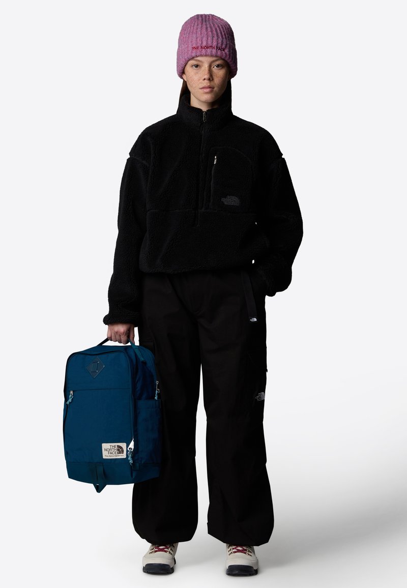 Pull en polaire noir avec fermeture éclair courte, pantalon cargo noir et un sac à dos bleu canard avec logo et accent en forme de diamant, porté avec des bottes claires.