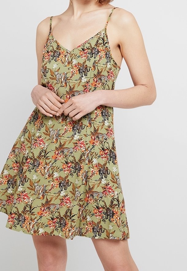 Femme portant une robe sans manches vert olive à motifs floraux et léopard, debout les mains jointes à la taille.