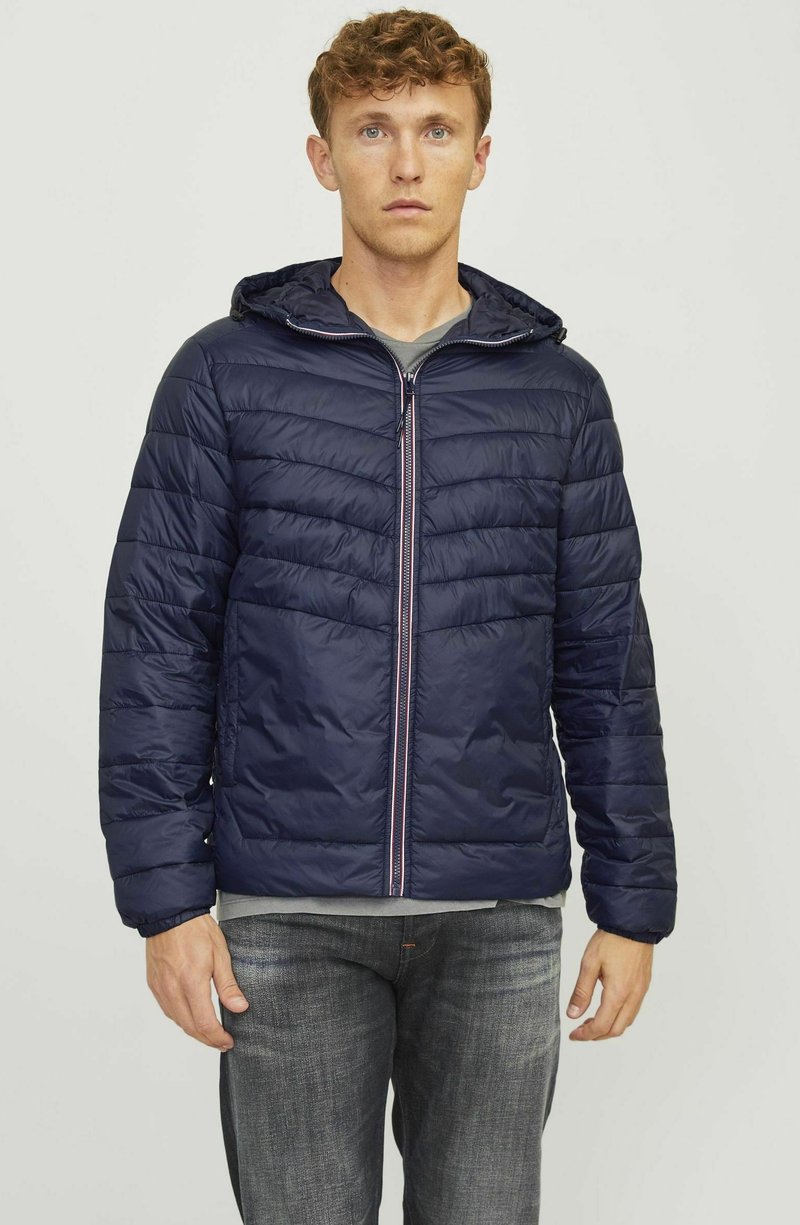 Chaqueta de plumas azul marino con cierre frontal, capucha y texturizado con acolchado en forma de chevrón. Presenta puños elásticos y un interior forrado. Llevada sobre una camiseta gris.