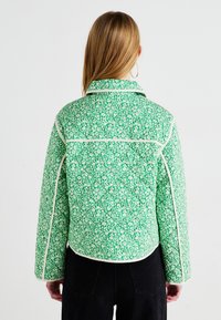 Giacca con motivo floreale verde e piping bianco, design imbottito, colletto e maniche lunghe. Indossata con pantaloni neri, vista da dietro.