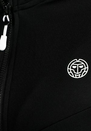 Schwarzer Stoff mit einem weißen Reißverschlussanhänger links und einem kleinen weißen runden Emblem mit einem stilisierten Gesicht rechts.