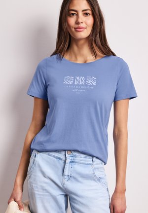 Femme portant un T-shirt bleu avec des carrés à motif animalier et le texte "La Vita Da Bohème", un jean délavé clair, et tenant un chapeau beige.