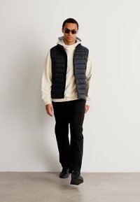 Μαύρη πούφφερ γιλέκα πάνω από μια ανοιχτή κρεμ φούτερ, συνδυασμένη με μαύρα παντελόνια και σκούρα sneakers. Το μοντέλο φορά γυαλιά ηλίου και στέκεται σε ένα ουδέτερο φόντο.