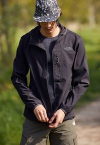 Czarna kurtka softshell z kapturem, zamykanymi kieszeniami i elastycznymi mankietami, zestawiona z szarym koszulką sportową i oliwkowymi spodniami cargo. Czapka w wzory.