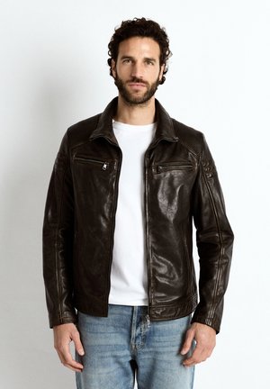 Man met krullend haar en baard, draagt een donkerbruine leren jas over een wit shirt en blauwe jeans, staand tegen een witte achtergrond.