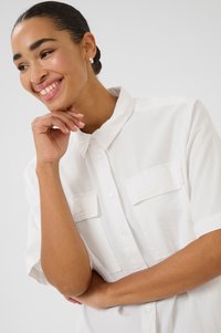 Chemise blanche à manches courtes avec boutonnage, dotée de deux poches poitrine, d'un col pointu et d'un tissu texturé. Surface lisse avec une coupe décontractée.