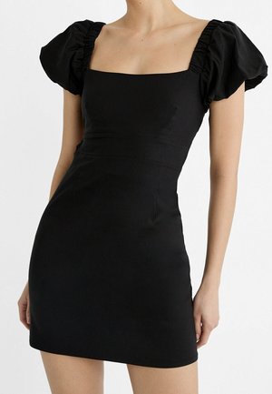 Kobieta w dopasowanej czarnej mini sukience z bufiastymi krótkimi rękawami i kwadratowym dekoltem na jednolitym białym tle.
