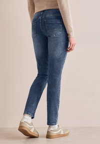 Jean en denim slim fit, bleu foncé, avec poches arrière, détails de couture visibles et texture lisse, portés avec des baskets beiges.