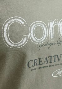 Oliwkowa koszulka z dużym białym i ciemnoszarym napisem, w tym "Core" i "Creative Dept", na miękkim, teksturowanym materiale.