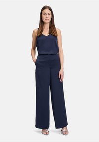 Débardeur en satin bleu marine avec un col en V, associé à un pantalon large bleu marine. Le mannequin se tient avec les mains dans les poches, portant des talons aiguilles argentés.