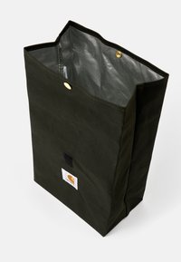 Canvasbag i mörkgrön färg med en strukturerad form, gyllene metallknapp som stängning och en logotypapplikation på sidan; insidan är fodrad med ett nätmaterial.