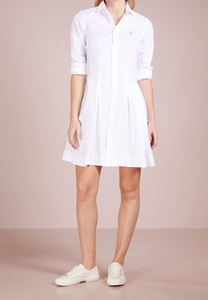 Biała sukienka na guziki z długimi rękawami podwiniętymi do łokci, z plisowaną spódnicą i małym wyszywanym logo na piersi. Nosi ją w białych trampkach.