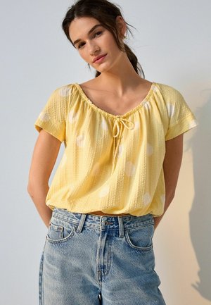 Jeune femme aux cheveux foncés portant un chemisier jaune à pois et un jean bleu clair, debout devant un fond clair uni.