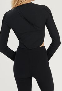 Schwarzes, geripptes Langarmoberteil mit einem ausgeschnittenen Rückendesign, raffinierten Schulternahtdetails und taillierter Silhouette. Kombiniert mit passenden schwarzen Hosen.