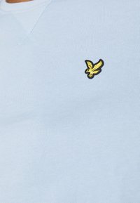 Világoskék kötött pulóver kerek nyakkal. Bal mellen egy kicsi fekete és sárga hímzett sas logo található.