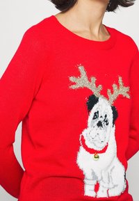 Pull rouge en tricot avec un motif de chien blanc, des accents noirs, des bois scintillants et une clochette dorée sur un collier rouge.