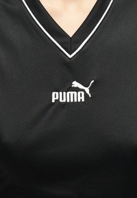 Μαύρο αθλητικό πουκάμισο με σχήμα V και λευκό λογότυπο Puma καθώς και λευκή διακόσμηση γύρω από το ντεκολτέ στο πάνω μέρος του στήθους ενός ατόμου.