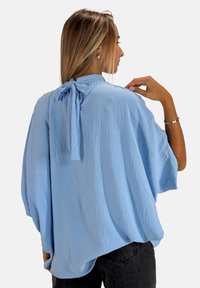 Lyseblå, krøllet bluse med afslappet pasform, der har en bundet halsudskæring bagpå og brede ærmer. Båret med sorte denim bukser.