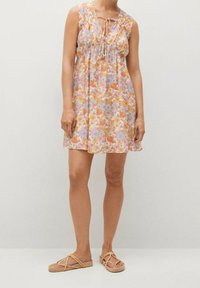 Robe sans manches à motifs floraux dans des couleurs pastel, avec une taille cintrée et un détail à nouer au niveau du décolleté. Associée à des sandales plates à lanières.