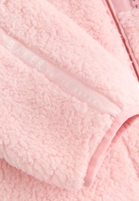 Blødt lyserødt sherpa fleece stof med en lomme med en lille hvid "BAKER by Ted Baker"-logo og en pink lynlås i nærheden.
