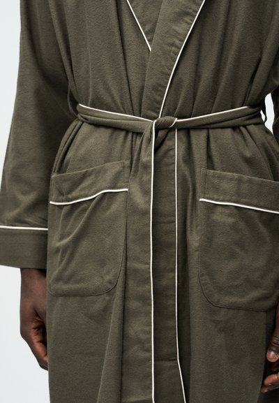 Ρόμπα ελιάς με λευκή επένδυση, μεγάλες μπροστινές τσέπες και δεμένη ζώνη στη μέση, φορεμένη από ένα άτομο.