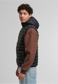 Vycpaná vesta s čiernym prešívaným povrchom a hnedými rukávmi z mikiny. Obsahuje kapucňu a žebrované manžety. Nosená cez svetlo modré džínsy.