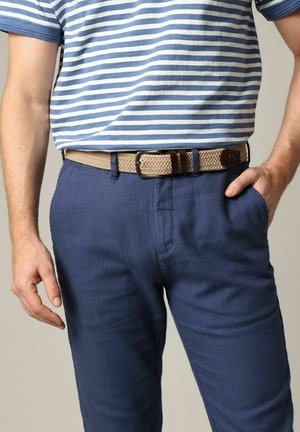 Homme portant une chemise rayée bleu et blanc rentrée dans un pantalon bleu avec une ceinture tissée beige, main droite dans la poche, main gauche détendue sur le côté.