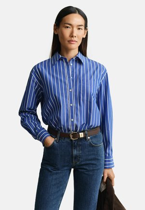 Frau mit langen dunklen Haaren, die ein blau-weiß gestreiftes Hemd mit Knöpfen trägt, in blaue Jeans mit braunem Gürtel gesteckt, und eine dunkle Jacke hält.